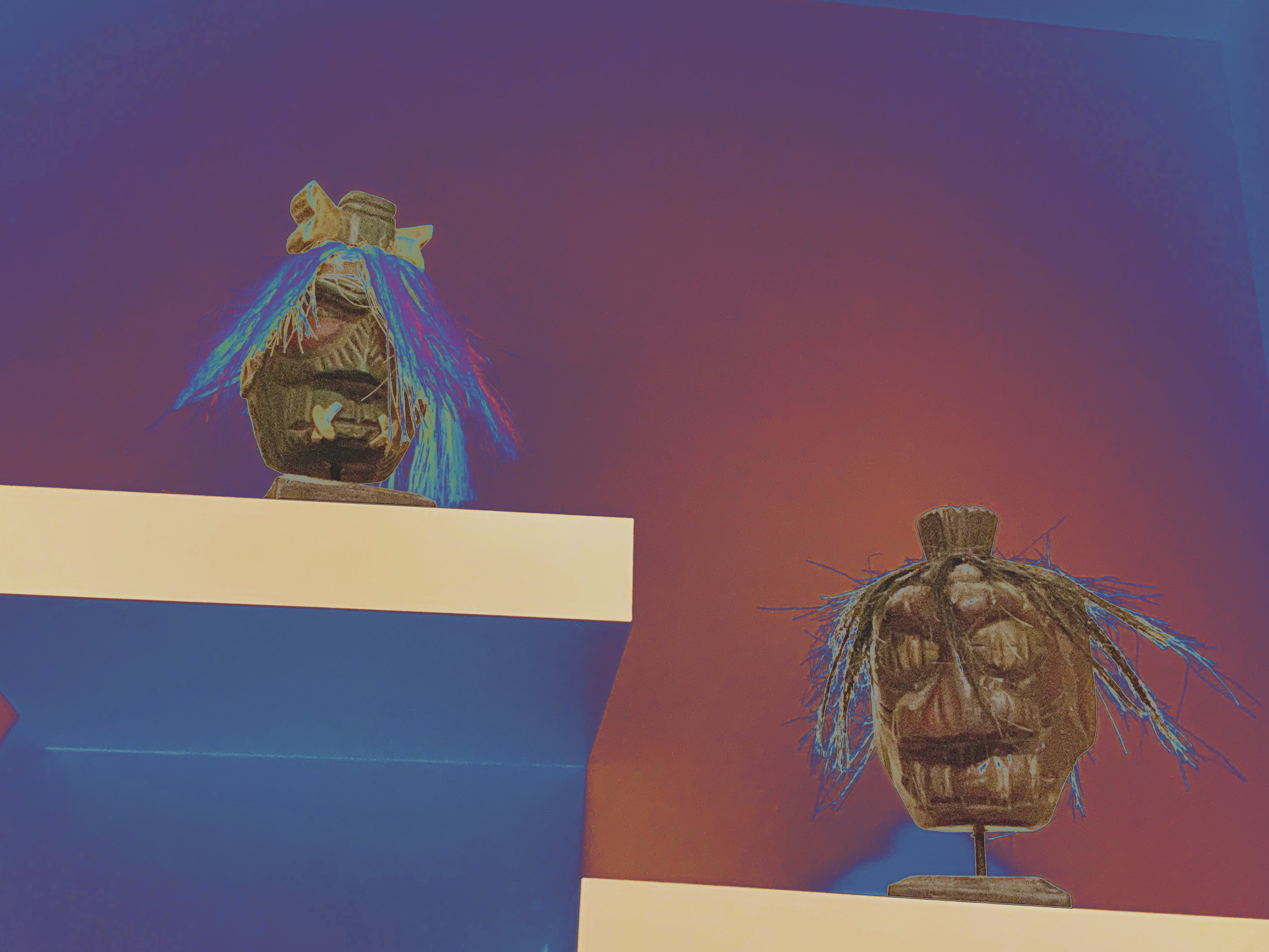 Two carved wooden heads with colorful fiber hair on white shelves, in a color-shifted photo